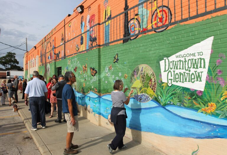 Community members admiring the newly-unveiled mural in downtown Glenview, a brightly-painted orange, green, and blue landscape featuring colorful wildlife, community life, and text that says "Welcome to Downtown Glenview."