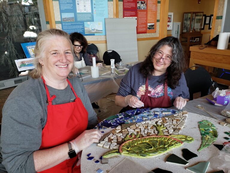 Two women seated at a table with aprons on constructing tile mosaics.