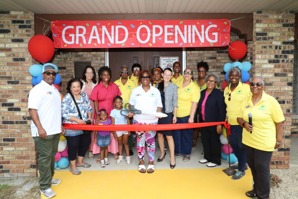 Riverdale Park Community Members stand in front of a large red banner that says "Grand Opening" holding scissors that will cut a long red ribbon to symbolize the completion of the project.